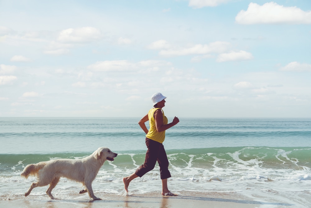 Happy,Elderly,Woman,Running,Along,A,Beach,With,Her,Golden